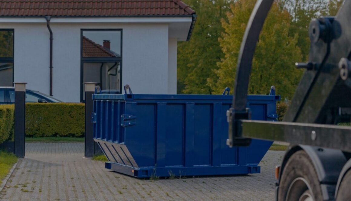 Blaue Absetzmulde steht aufstellbereit in einer gepflasterten Hofeinfahrt, daneben die offene Durchfahrt mit Torpfosten