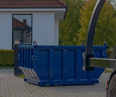 Blaue Absetzmulde steht aufstellbereit in einer gepflasterten Hofeinfahrt, daneben die offene Durchfahrt mit Torpfosten